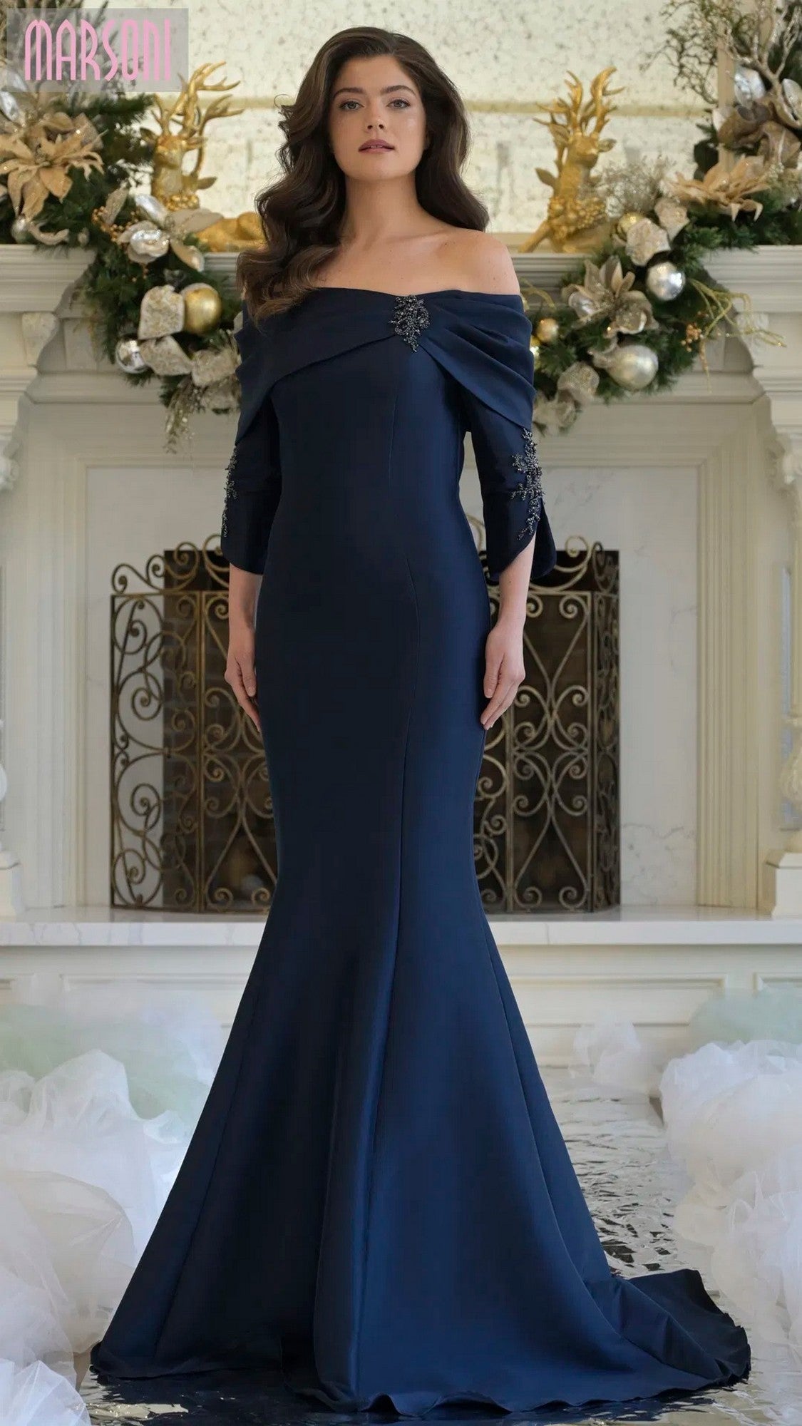 A woman in a navy Marsoni MV1360 long formal dress stands before a festive fireplace decorated with holiday ornaments and greenery.